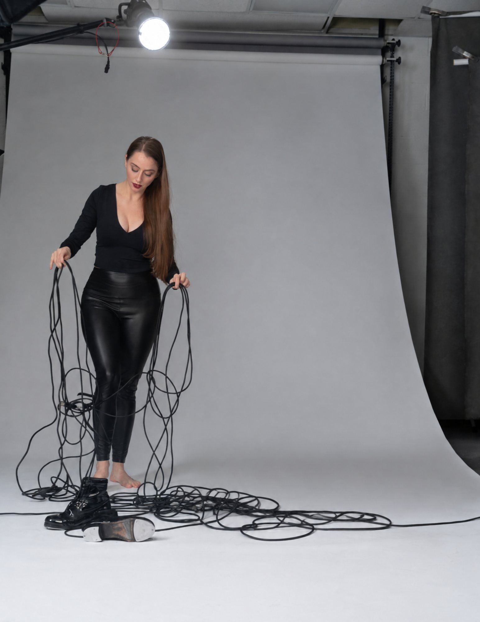 Hillary-Marie in a photo studio, dressed in black, holding up loops of long black cables with a pair of tap shoes at her feet