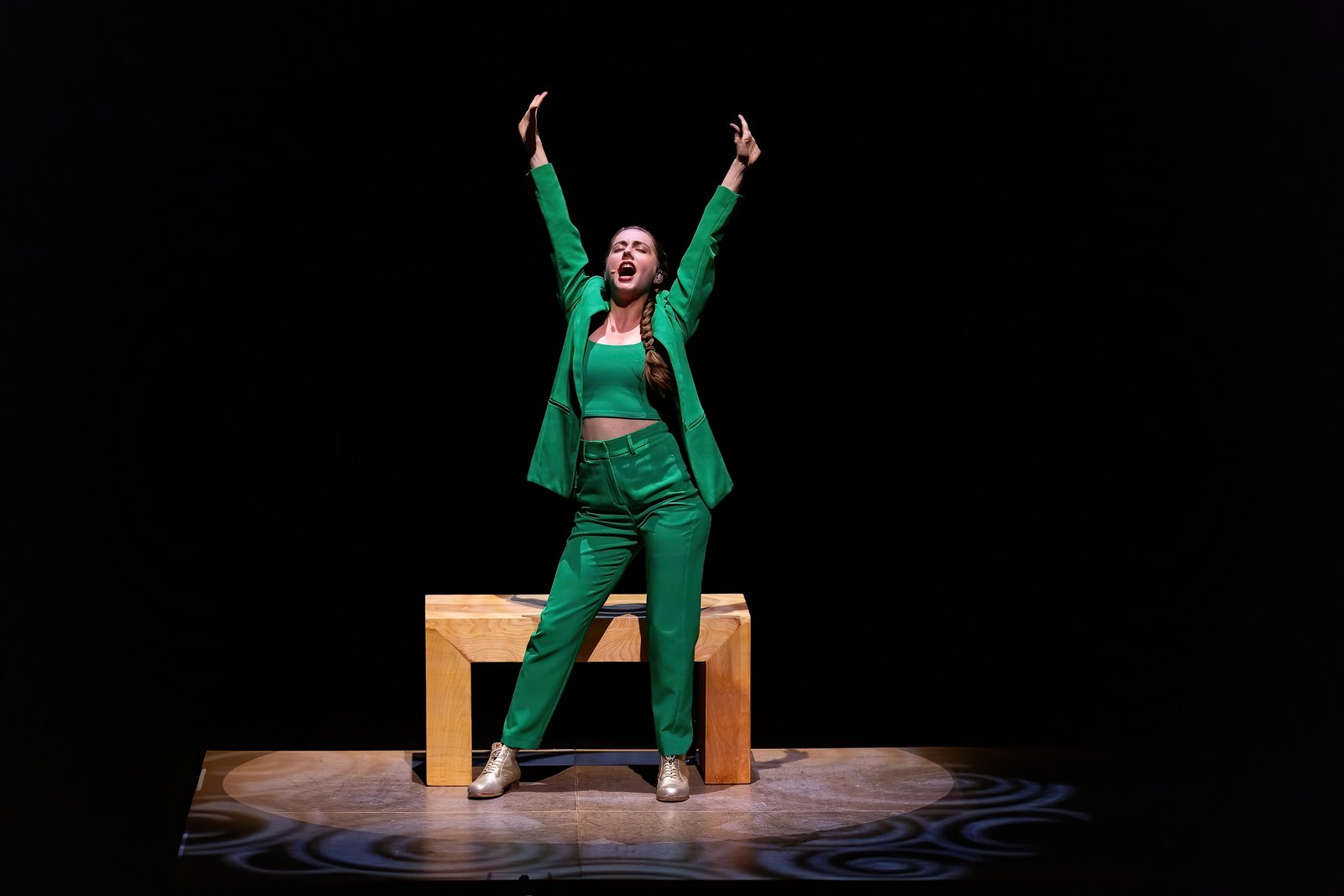 Hillary-Marie on stage in In The Pocket, in a green suit and gold tap shoes, arms thrown up mid-shout in front of a wooden bench